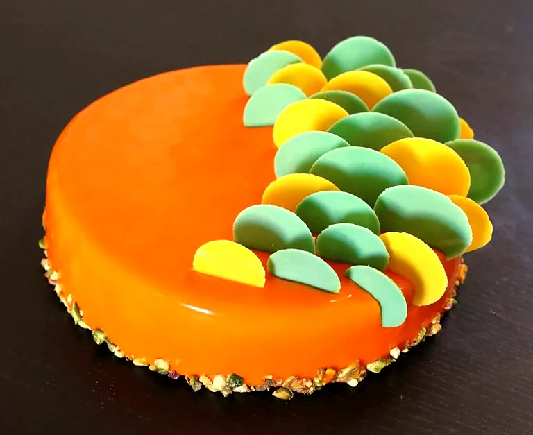 Entremets au glaçage orange, décor de demi-disques verts et jaunes, pied de pistaches concassées
